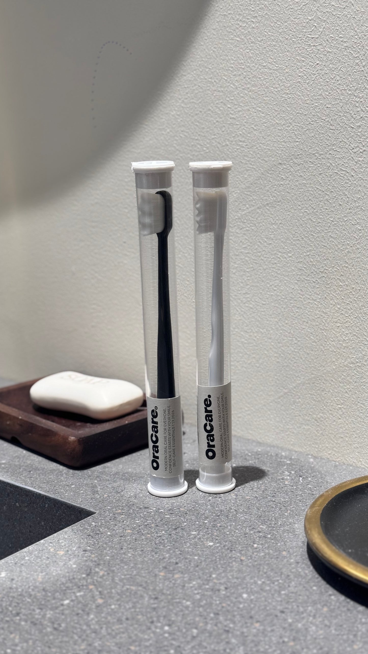 Two OraCare toothbrushes in clear cylindrical tubes with white caps, standing on a gray countertop against a textured off-white wall. A soap dish with a bar of soap is visible on the left.